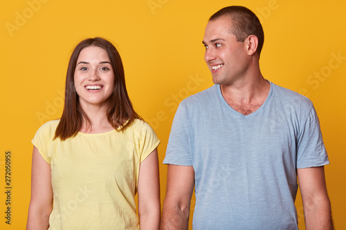Fotografie Beautiful couple, handsome man looking at her girlfriend and smiling while standing over yellow background, charming female looking directly at camera with toothy smile
