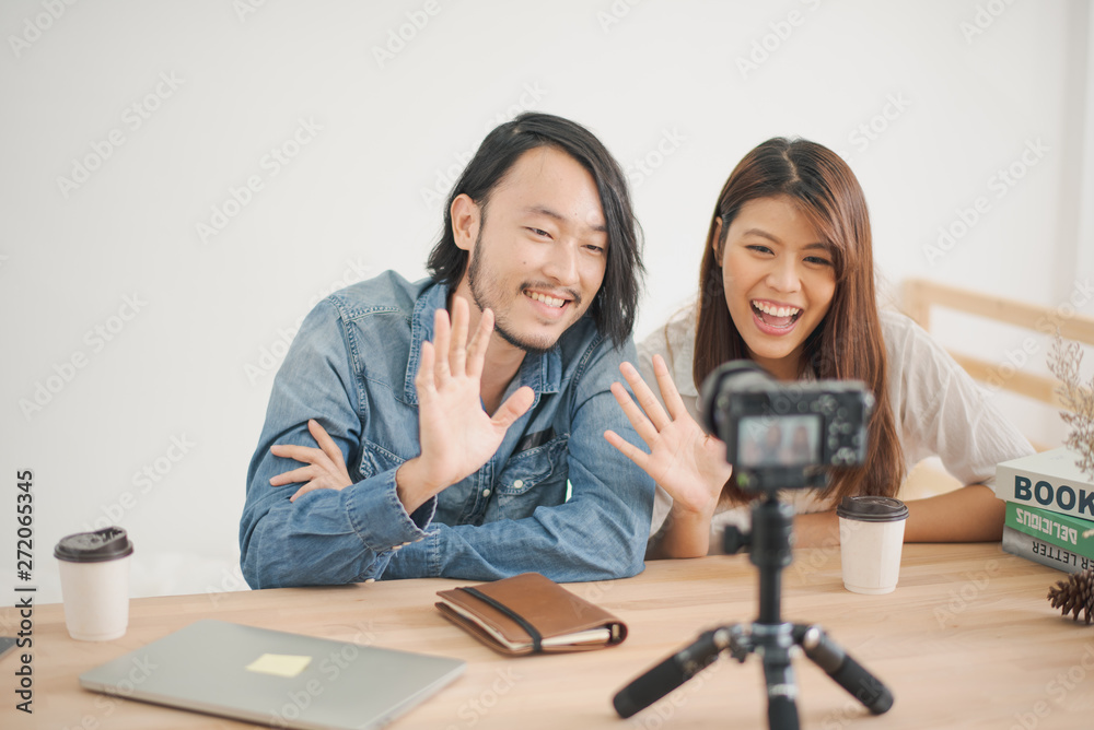 Young Asian man and woman are waving the hand with the camera which standing on the wood table. the couple man and woman sitting in the home and record video clip. blogger and vlog concept Young Asian man and woman are waving the hand with the camera which standing on the wood table. the couple man and woman sitting in the home and record video clip. blogger and vlog concept