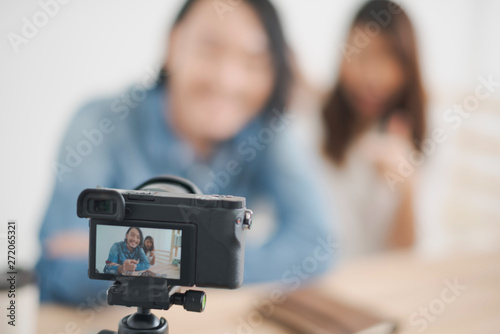 Focus at camera, Young Asian man and woman are talking with the camera which standing on the wood table. the couple man and woman sitting in the home and record video clip. blogger and vlog concept.