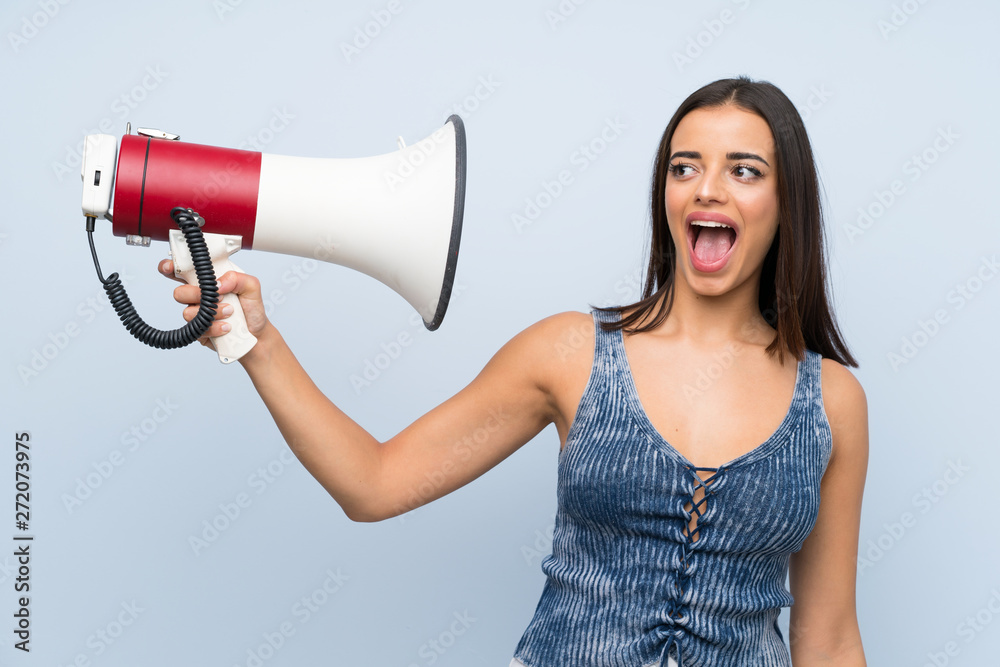 Naklejka premium Young woman over isolated blue wall holding a megaphone