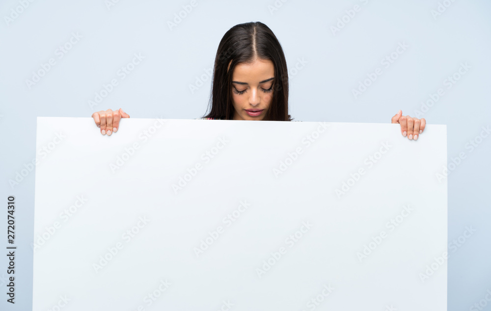 Young woman over isolated blue wall holding an empty white placard for insert a concept Young woman over isolated blue wall holding an empty white placard for insert a concept