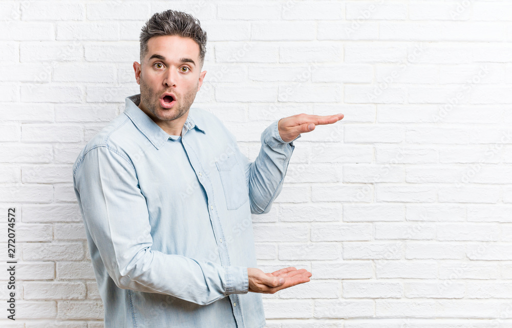 Young handsome man against a bricks wall shocked and amazed holding a copy space between hands. Young handsome man against a bricks wall shocked and amazed holding a copy space between hands.