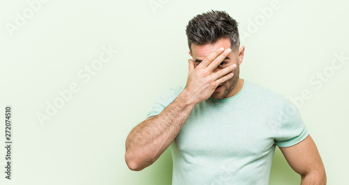Young handsome man against a green background blink at the camera through fingers, embarrassed covering face.