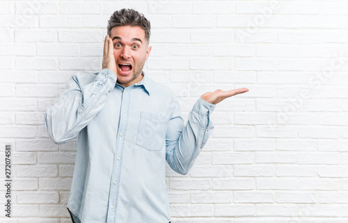 Young handsome man against a bricks wall holds copy space on a palm, keep hand over cheek. Amazed and delighted.