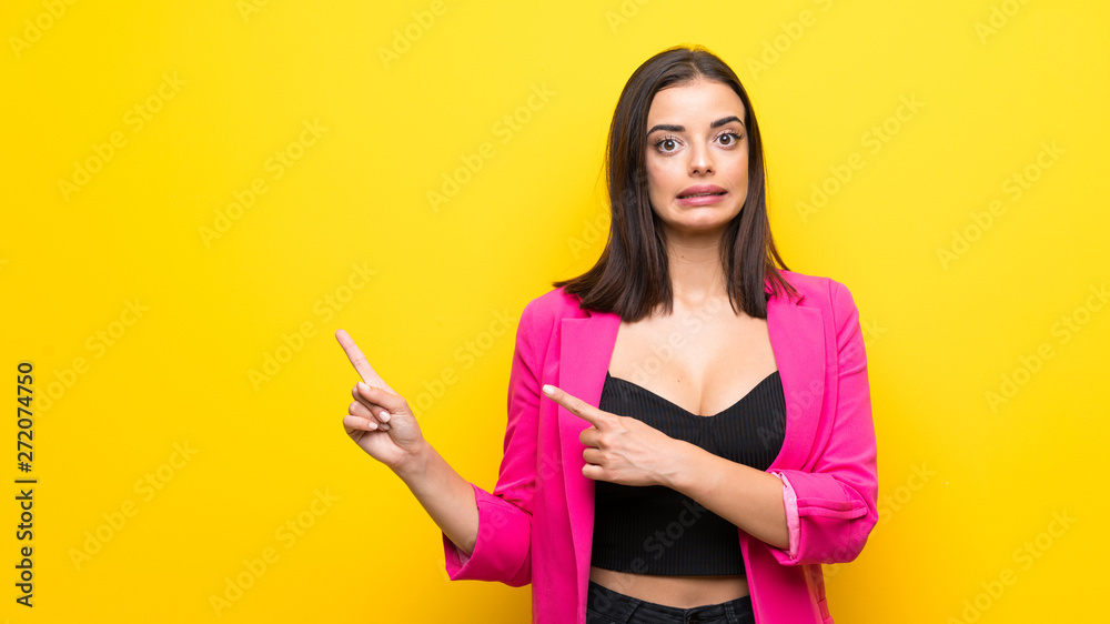 Young woman over isolated yellow background frightened and pointing to the side Young woman over isolated yellow background frightened and pointing to the side