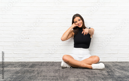 Pretty young woman sitting on the floor making phone gesture and pointing front