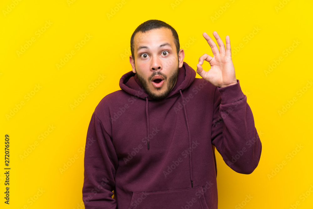 Colombian man with sweatshirt over yellow wall surprised and showing ok sign Colombian man with sweatshirt over yellow wall surprised and showing ok sign