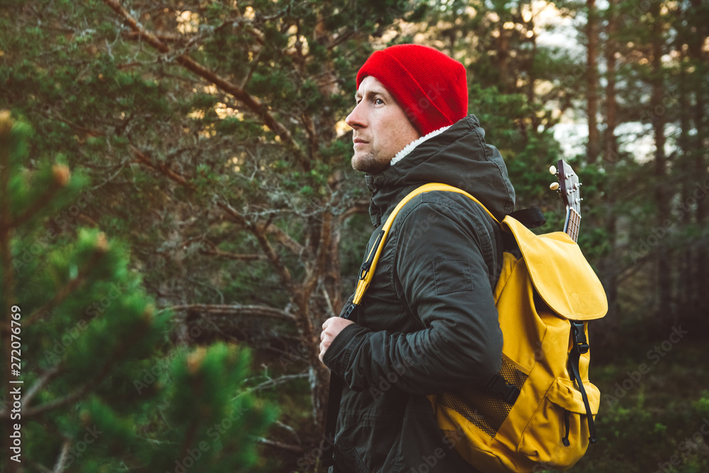 Traveler man is standing on a trail in the middle of a forest with a guitar. Wearing a yellow backpack in a red hat. Lifestyle Travel Concept. Shoot from the back