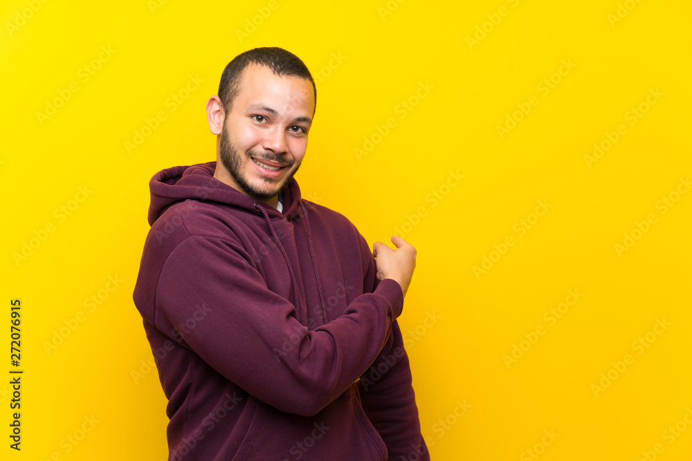 Colombian man with sweatshirt over yellow wall pointing back Colombian man with sweatshirt over yellow wall pointing back