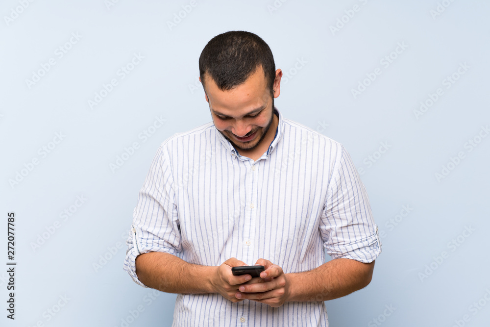 Colombian man over isolated blue wall sending a message with the mobile Colombian man over isolated blue wall sending a message with the mobile