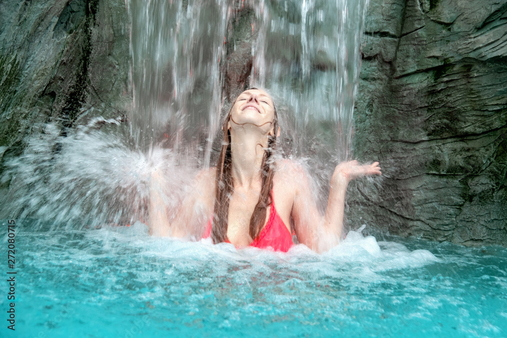 young sexy blond woman in fuchsia bikini raises her hands against the splashing water of the waterfall in the swimming pool facility