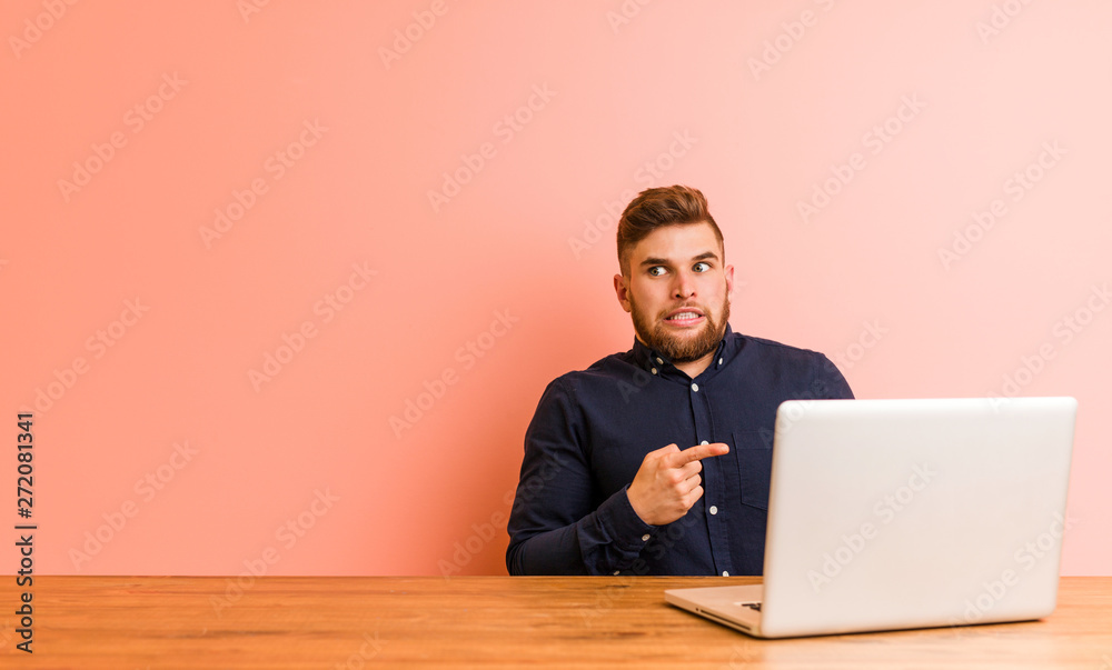 Young man working with his laptop shocked pointing with index fingers to a copy space. Young man working with his laptop shocked pointing with index fingers to a copy space.