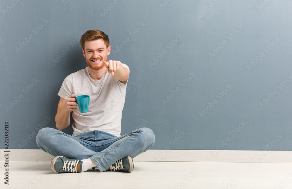 Young redhead student man sitting on the floor cheerful and smiling pointing to front. He is holding a coffee mug. Young redhead student man sitting on the floor cheerful and smiling pointing to front. He is holding a coffee mug.