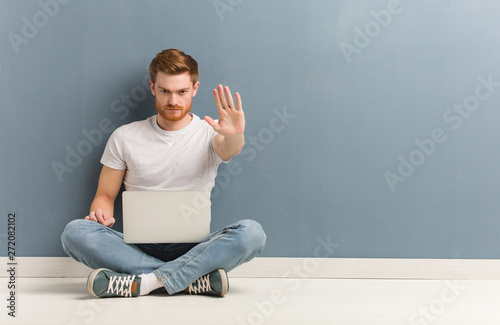 Young redhead student man sitting on the floor putting hand in front. He is holding a laptop.