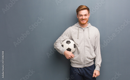 Young redhead fitness man cheerful with a big smile. He is holding a soccer ball. © Asier
