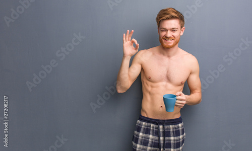 Young shirtless redhead man cheerful and confident doing ok gesture. He is holding a coffee mug.