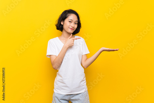 Asian young woman over isolated yellow wall holding copyspace imaginary on the palm to insert an ad