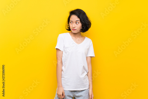Asian young woman over isolated yellow wall making doubts gesture looking side