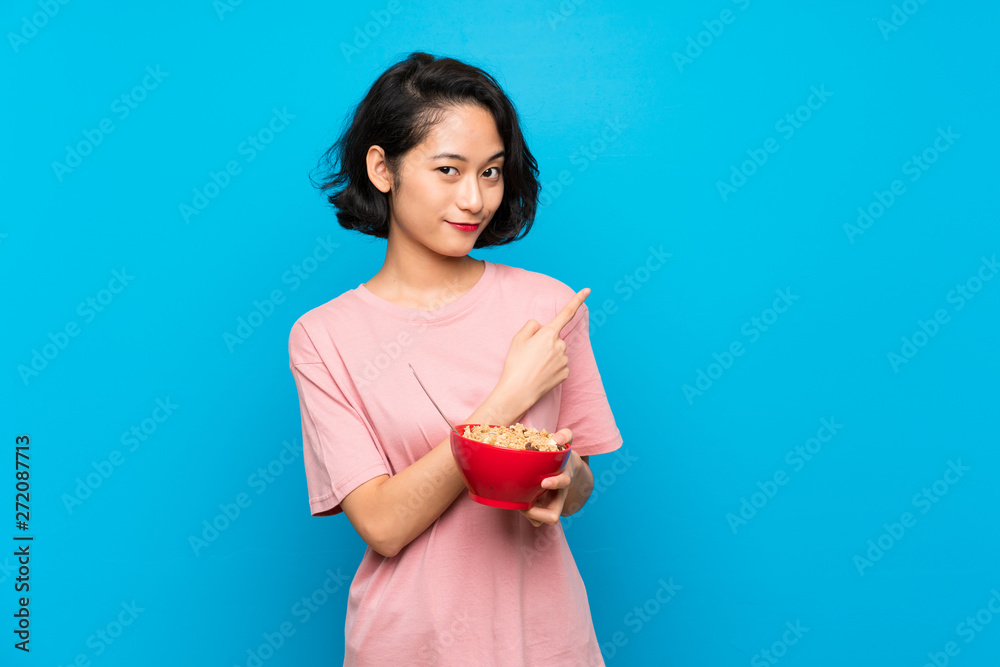 Asian young woman holding a bowl of cereals pointing to the side to present a product Asian young woman holding a bowl of cereals pointing to the side to present a product