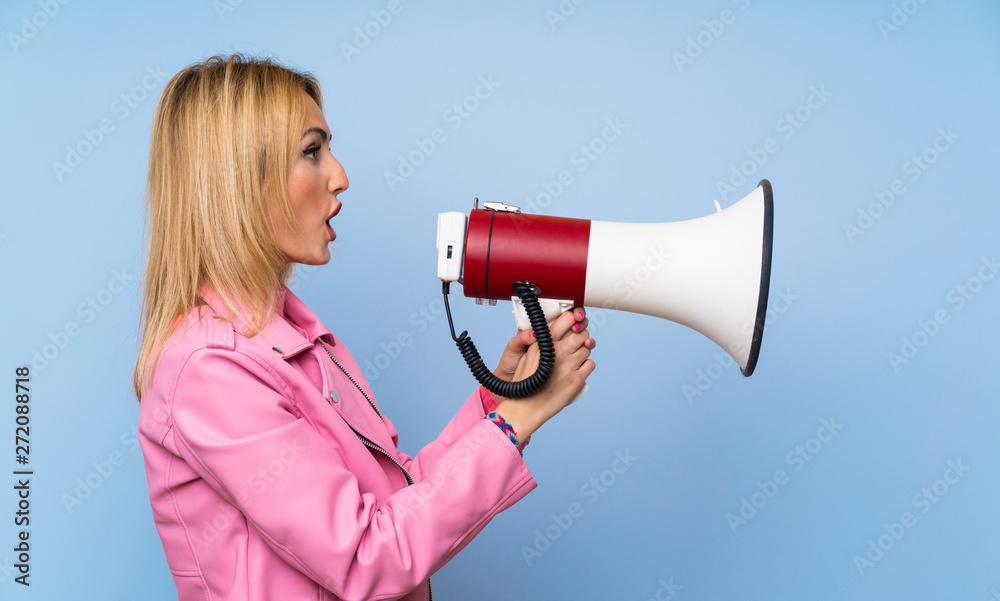 Young blonde woman with pink jacket over isolated blue background shouting through a megaphone Young blonde woman with pink jacket over isolated blue background shouting through a megaphone