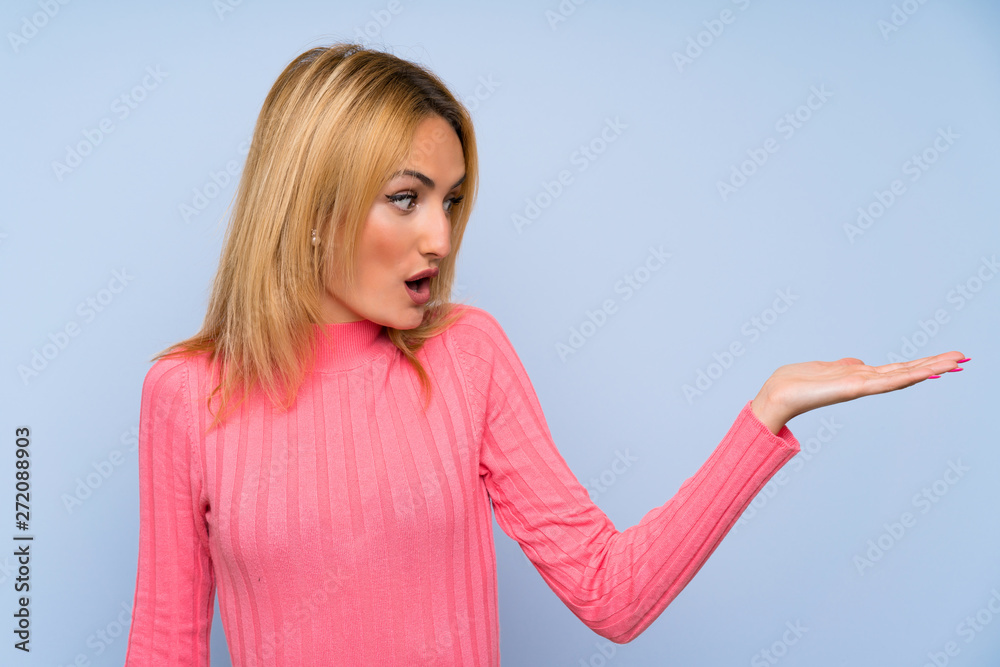 Fototapeta premium Young blonde woman with pink sweater over isolated blue background holding copyspace imaginary on the palm