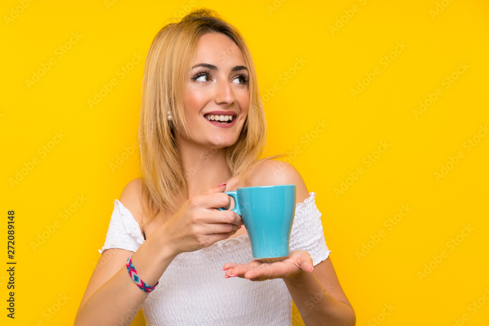 Young blonde woman over isolated yellow wall holding hot cup of coffee Young blonde woman over isolated yellow wall holding hot cup of coffee
