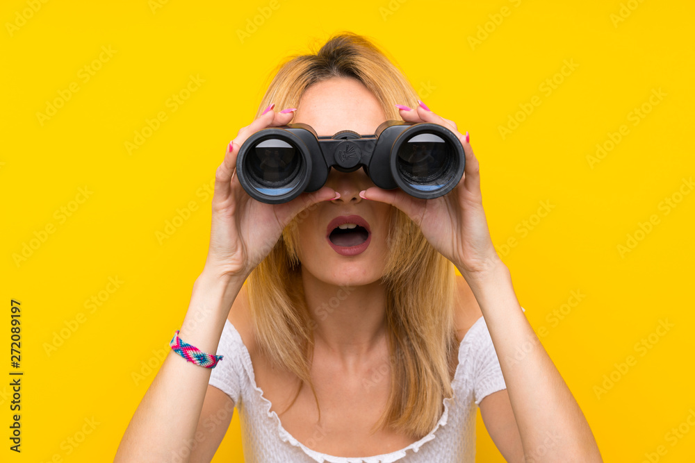 Young blonde woman over isolated yellow wall with black binoculars