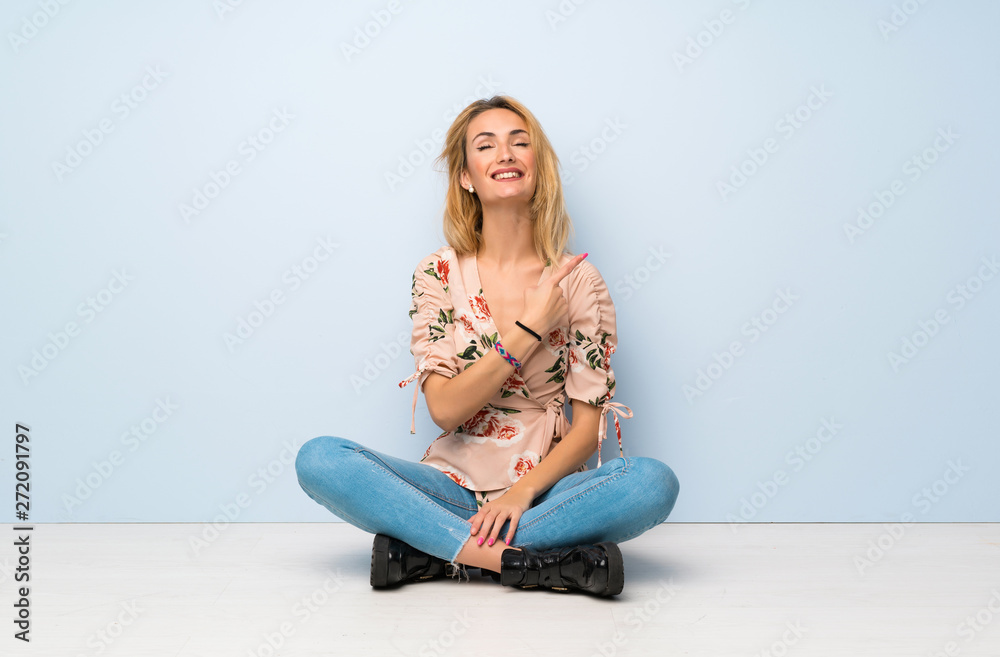Young blonde woman sitting on the floor pointing to the side to present a product Young blonde woman sitting on the floor pointing to the side to present a product