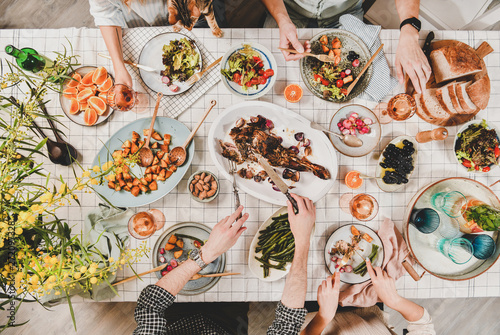 Family or friends gathering dinner. Flat-lay of peoples hands with roasted lamb shoulder, salads, vegetables, wine, mimosa branches over white checkered tablecloth, top view. Celebration party dinner