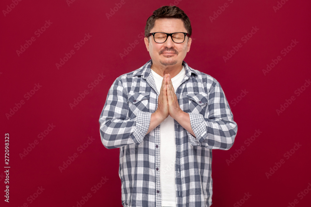 Portrait of relaxed handsome middle aged business man in casual checkered shirt and eyeglasses standing with palm hands and doing yoga meditating. indoor studio shot, isolated on dark red background. Portrait of relaxed handsome middle aged business man in casual checkered shirt and eyeglasses standing with palm hands and doing yoga meditating. indoor studio shot, isolated on dark red background.
