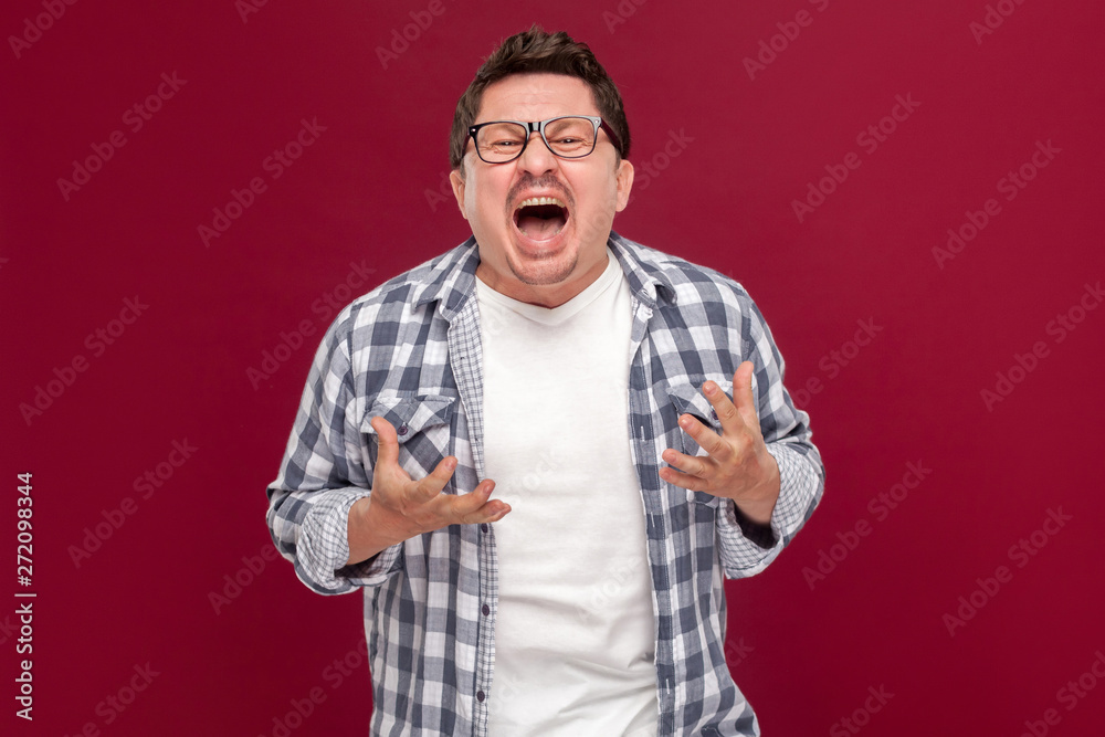 Portrait of handsome middle aged business man in casual checkered shirt and eyeglasses standing, raised arms, looking at camera and screaming. indoor studio shot, isolated on dark red background. Portrait of handsome middle aged business man in casual checkered shirt and eyeglasses standing, raised arms, looking at camera and screaming. indoor studio shot, isolated on dark red background.