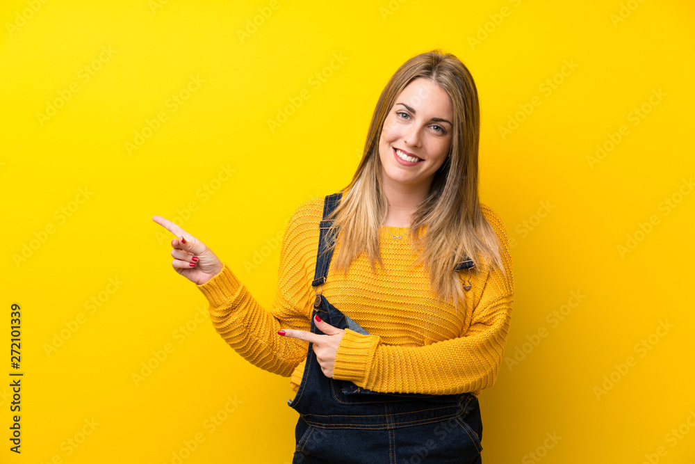 Woman with overalls over isolated yellow wall pointing finger to the side Woman with overalls over isolated yellow wall pointing finger to the side