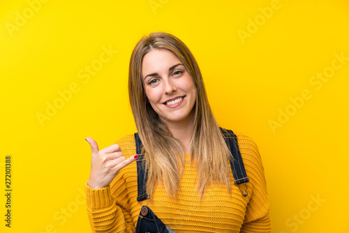 Woman with overalls over isolated yellow wall making phone gesture