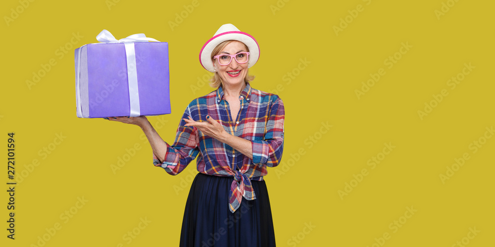 Fototapeta premium Want present? Happy beautiful modern grandma in white hat and in checkered shirt standing, holding big gift box with toothy smile, pointing hand and looking at camera. Studio shot, yellow background