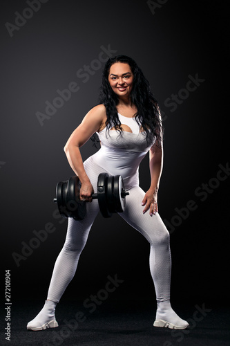 Size plus woman sporty fit woman in white sportswear, athlete with dumbbells makes fitness exercising on black background with lights. Motivation for fat people.