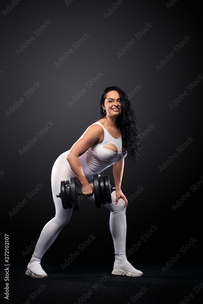 Size plus woman sporty fit woman in white sportswear, athlete with dumbbells makes fitness exercising on black background with lights. Motivation for fat people. Size plus woman sporty fit woman in white sportswear, athlete with dumbbells makes fitness exercising on black background with lights. Motivation for fat people.