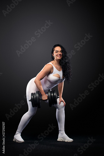 Size plus woman sporty fit woman in white sportswear, athlete with dumbbells makes fitness exercising on black background with lights. Motivation for fat people.