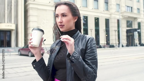 beautiful young girl in a black leather jacket nibbling on sunglasses against the background of a city street. drinks coffee from a paper Cup.