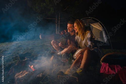 Young couple baking sausages on the campfire and drinking beer in the forest hill in the dusk