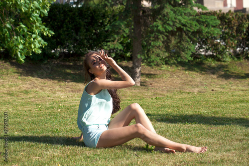 Young brunette woman in a turquoise blouse posing sitting on a green lawn in a summer park