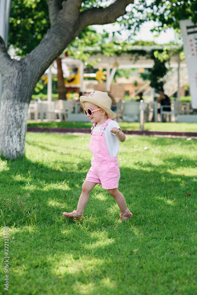 Stylish girl in a pink overalls, hat and pink glasses walks on the green grass. Summer vacation concept. Stylish girl in a pink overalls, hat and pink glasses walks on the green grass. Summer vacation concept.