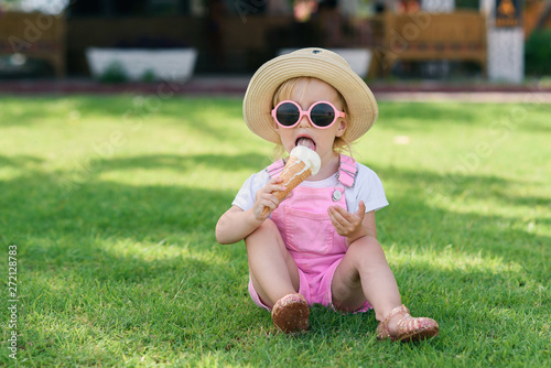 Bild auf Leinwand Stylish toddler girl in a hat and pink sunglasses eats with pleasure ice cream on a green lawn on a hot sunny day