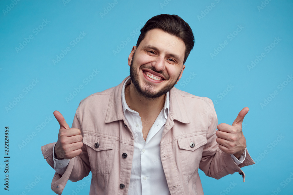 Headshot of handsome young male wearing pink stylish jeans jacket, smiling and showing thumb up over blue background. Headshot of handsome young male wearing pink stylish jeans jacket, smiling and showing thumb up over blue background.