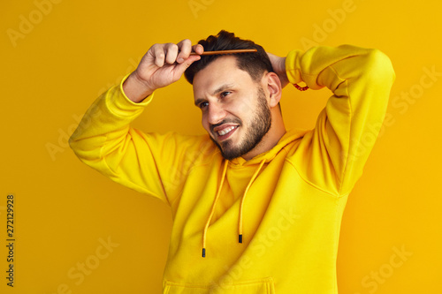 Attractive young boy, wearing yellow hoodie, smiling and combing his hair, makes hairstyle by himself, isolated over yellow wall © Davidovici