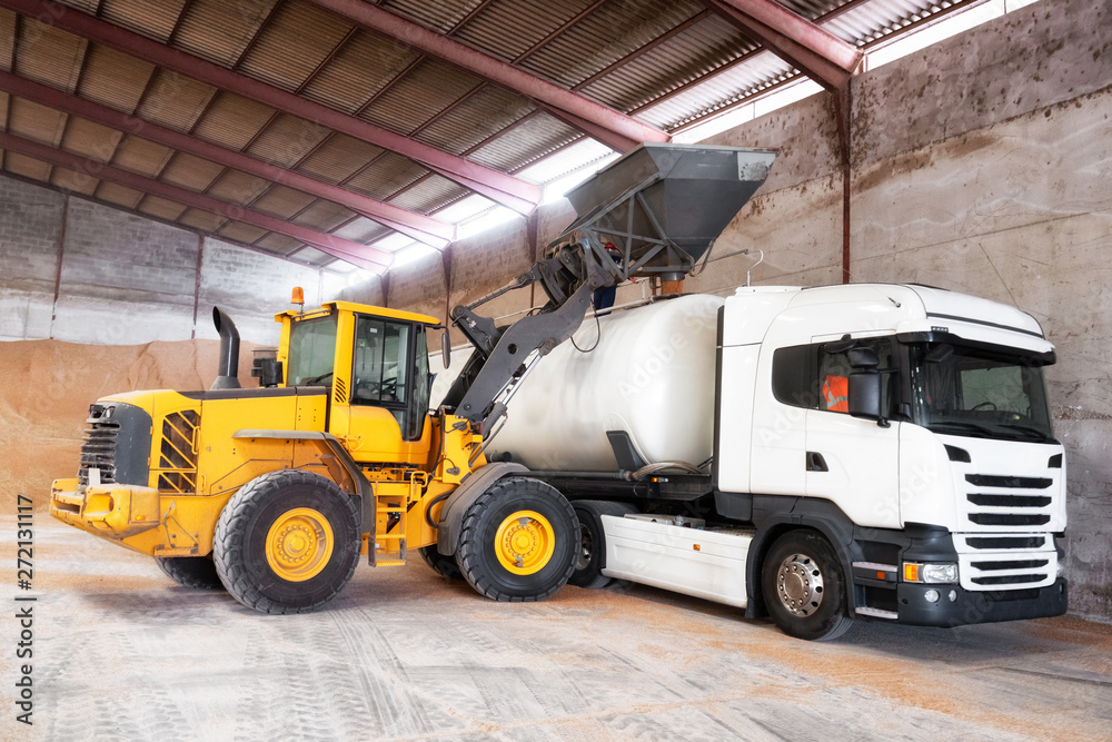 Tractor loading grain into truck at at grain processing plant warehouse, during harvesting time .