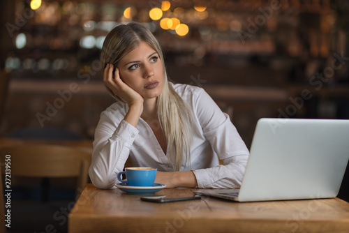 Young pensive businesswoman using computer in cafeteria and looking away