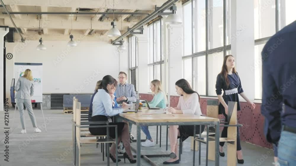 Business team holds a meeting at a large table. An employee gets up and goes to colleagues for a glass presentation board. Coworking. Loft style office space Business team holds a meeting at a large table. An employee gets up and goes to colleagues for a glass presentation board. Coworking. Loft style office space