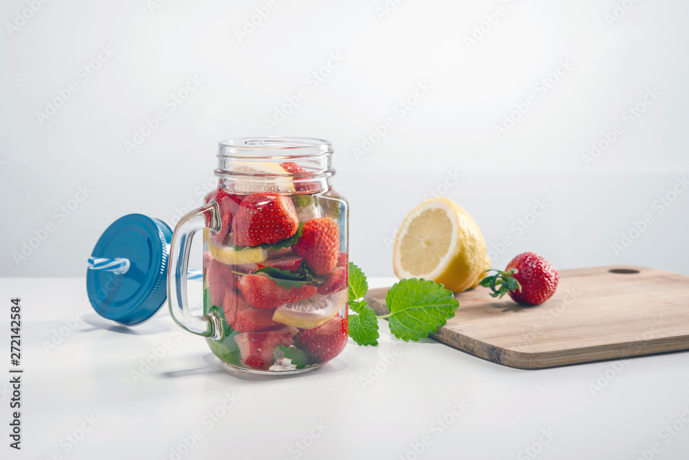 Mug of fruit water with strawberries, mint and lemon, stand on a wooden stand next to fresh strawberries, mint leaf, sliced lemon and a cap with a straw for drinking. Mug of fruit water with strawberries, mint and lemon, stand on a wooden stand next to fresh strawberries, mint leaf, sliced lemon and a cap with a straw for drinking.
