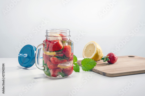 Mug of fruit water with strawberries, mint and lemon, stand on a wooden stand next to fresh strawberries, mint leaf, sliced lemon and a cap with a straw for drinking.