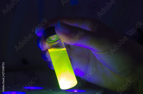 Researcher holding green photochemical reaction in glass vial under UV light in a dark chemistry laboratory